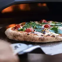 Wood-fired pizza oven with fresh pizza featuring greens and toppings, enhancing outdoor entertainment spaces