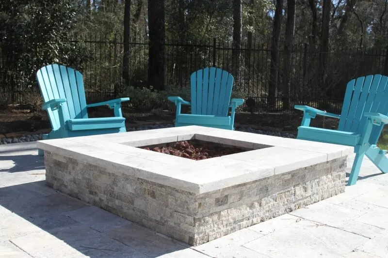 Modern stone fire pit with seating area featuring turquoise Adirondack chairs in luxury backyard design