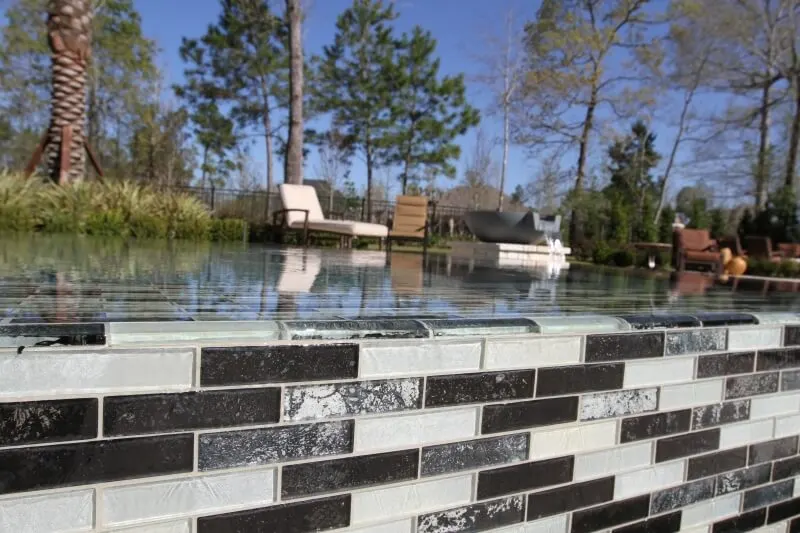 Glass tile negative edge luxury pool with waterfront backdrop and pine trees