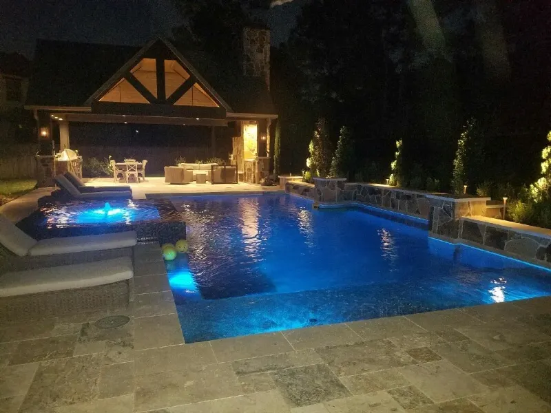 Luxury backyard pool with blue LED lighting, spa, and stone patio entertainment area at night