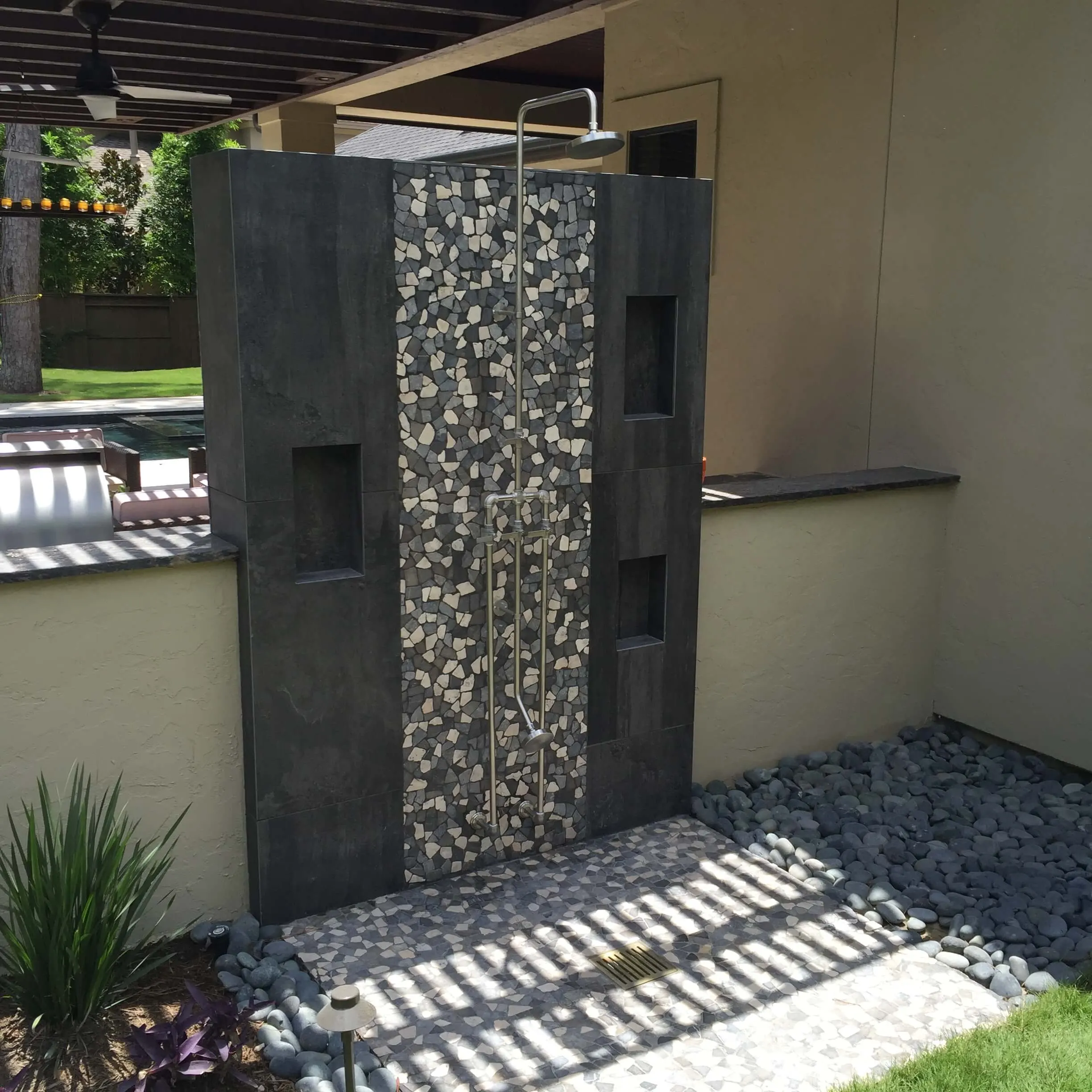 Modern outdoor shower with dark tile surround and pebble accent wall in luxury backyard space