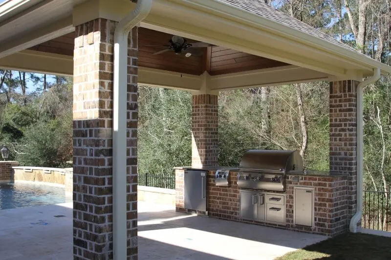 Custom outdoor kitchen with grill under brick-columned covered patio overlooking luxury backyard pool